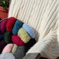 A cozy white, soft luxurious Cabled Afghan from Plymouth Yarn Co drapes over the back of a chair. Beside it, a wicker basket holds assorted colorful balls of yarn in red, green, blue, pink, and gray hues. A potted plant in the background adds a touch of greenery—perfect for cool fall or winter evenings.