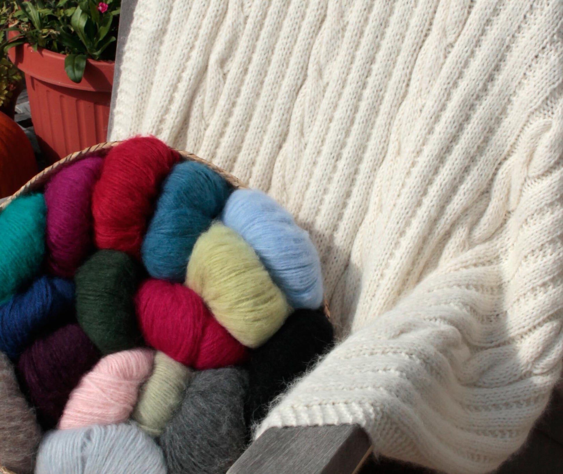 A cozy white, soft luxurious Cabled Afghan from Plymouth Yarn Co drapes over the back of a chair. Beside it, a wicker basket holds assorted colorful balls of yarn in red, green, blue, pink, and gray hues. A potted plant in the background adds a touch of greenery—perfect for cool fall or winter evenings.