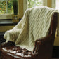 A cozy, cream-colored Cabled Afghan by Plymouth Yarn Co is draped over a brown leather armchair with stud detailing, positioned next to a window. Green plants, visible through the window and on the windowsill, add a touch of nature to the warm interior setting—perfect for cool fall or winter days.