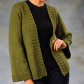 A person with light skin is stylishly dressed in a Woman's Kimono Sweater from Plymouth Yarn Co, knitted in a vibrant green worsted weight yarn. They’re wearing it over a black shirt and dark jeans. With one hand, they hold the sweater open, revealing a silver ring on their finger, against a plain, neutral background.