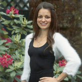 A woman with long dark hair, dressed in a black dress and a white Ladies Shrug Pattern cardigan from Plymouth Yarn Co, stands in front of pink flowering plants outdoors. She is smiling with her right hand resting on her hip, capturing the essence of holiday wear.