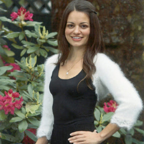 A woman with long brown hair is smiling while standing outdoors with one hand on her hip. She is wearing a black dress and a white fuzzy shrug from Plymouth Yarn Co, made from an exquisite angora blend. Behind her, there are lush green plants with pink flowers, creating the perfect backdrop for holiday wear.