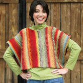 A person stands smiling with hands on hips, showcasing the Side to Side Vest by Plymouth Yarn Co, which is crafted from variegated yarns. The background features wooden doors, highlighting the cozy, artisanal vibe.