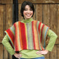 A person with short dark hair smiles at the camera. They are wearing the vibrant, striped "Side to Side Vest" from Plymouth Yarn Co over a green turtleneck shirt and blue jeans. They stand in front of a wooden door with their hands on their hips.