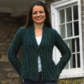 A woman with long brown hair smiles while standing outdoors. She is wearing a Plymouth Yarn Co Women's Super Bulky Cabled Cardigan in green over a white top, with her hands in the pockets of her blue jeans. A stone wall and a white-framed window are visible in the background.