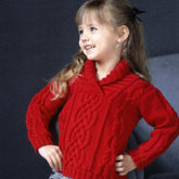 A young child with long blonde hair, tied with a black bow, smiles confidently while standing. She is wearing the vibrant red Kid's Shawl Collar Cabled Pullover by Plymouth Yarn Co and grey and white striped pants. The background is dark and blurred, emphasizing the girl.