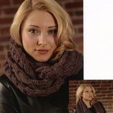 A woman with blonde hair is seen against a brick wall background, styled in a chic black jacket and the Plymouth Yarn Co Infinity Cowl, a brown chunky knitted accessory. She wears a subtle smile. An inset image shows her in the same outfit, looking to the side. The Plymouth Yarn Co Infinity Cowl makes for an effortlessly charming and fun addition to her ensemble.