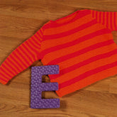 A vibrant Child's Pullover from Plymouth Yarn Co, featuring striking red and orange stripes, is laid flat on a wooden floor. Beside the knit pullover lies a large foam letter "E" in neon purple with a wavy pattern, enhancing the colorful display.