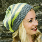 A smiling individual wearing a Plymouth Yarn Co Multi Stripe Slouch Hat for the Family in green and gray stripes stands against a stone wall. The person has long, blonde hair, and the beanie features a simple pattern that elegantly slouches back.