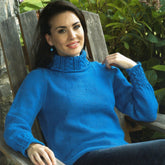 A person with long dark hair is sitting on a wooden chair outdoors, wearing the Women's Cable Detail Pullover by Plymouth Yarn Co in bright blue. The intricate cable details on the collar and cuffs add elegance to the outfit as green plants in the background contribute to a serene, natural setting.