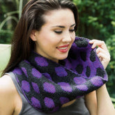 A woman with long dark hair is wearing a sleeveless gray top and a Double Knit Polka Dot Cowl by Plymouth Yarn Co. She is smiling and looking downwards, outdoors with greenery in the background.