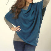 A woman stands with one hand on her hip, showcasing a teal Women's Poncho from Plymouth Yarn Co. The poncho, made from Worsted Merino Superwash yarn, is draped elegantly over her gray long-sleeve top and dark blue jeans. It features a lace trim along one edge, adding a touch of sophistication. Her long, wavy brown hair partially covers her face.