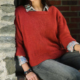 A person leans against a stone wall, dressed in a loose-fitting Plymouth Yarn Co Women's Dolman Pullover in red, layered over a plaid shirt and worn-in jeans. The person sports a colorful beaded bracelet on their left wrist, with their face out of the frame.