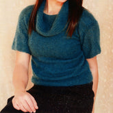 A person showcases a casually sophisticated look in a teal, short-sleeved cowl neck pullover from the Woman's Short Sleeve Cowl Neck Pullover Pattern by Plymouth Yarn Co, paired with a black skirt. The backdrop is a light-colored wall with subtle patterns. Their head is slightly tilted, and they have long, dark hair.