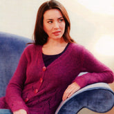 A woman with long brown hair sits on a blue velvet chair in what seems to be a cozy home or office setting. She is wearing the Women's Zig Zag Cardigan by Plymouth Yarn Co, a deep pink lacy sweater with large buttons, over a dark purple top. The background is softly lit, featuring warm, neutral tones. She looks slightly to the side with a calm expression.