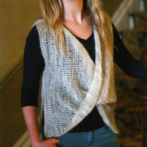 A person with long hair is pictured from the shoulders down, wearing a Plymouth Yarn Co Crochet Moebius-Look Vest over a black top. The outfit accessory showcases a loose, draped front reminiscent of its namesake style. The background appears to be an indoor staircase, slightly out of focus.