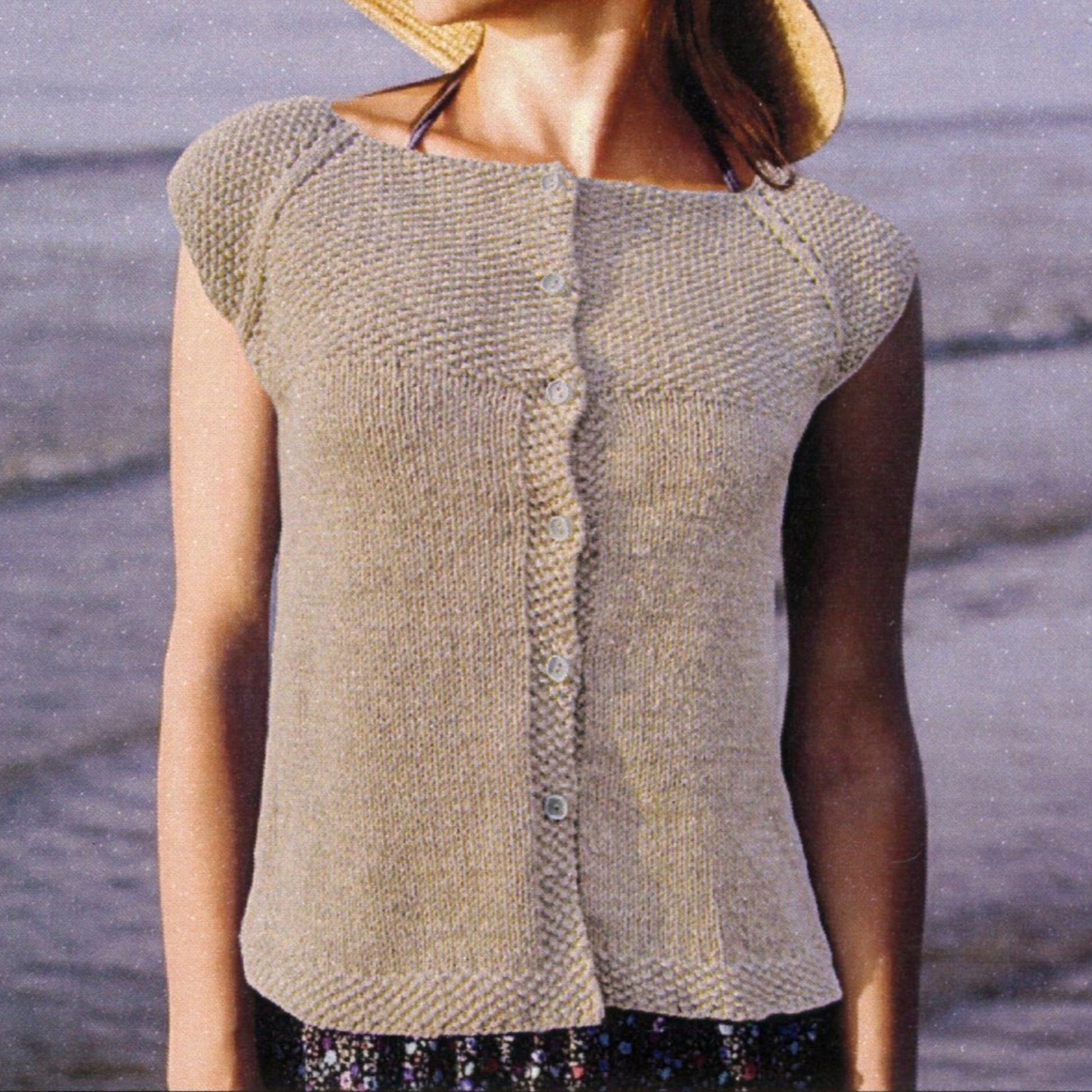 A person wearing the Seed Stitch Yoke Cardie by Knit One, Crochet Too stands on a beach. They are also sporting a wide-brimmed hat and a patterned skirt. The ocean is visible in the background.