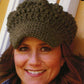 A woman is smiling while wearing a green Newsie Cap from Knit One, Crochet Too made from worsted weight yarn. The cap, which features a textured, chunky design and covers her forehead, is perfect for cozy indoor settings. Behind her, a partial white door frame and brown wall are visible.