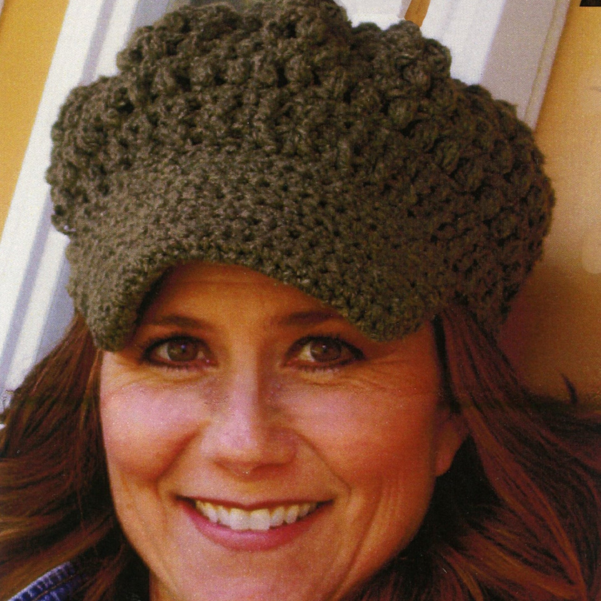 A woman is smiling while wearing a green Newsie Cap from Knit One, Crochet Too made from worsted weight yarn. The cap, which features a textured, chunky design and covers her forehead, is perfect for cozy indoor settings. Behind her, a partial white door frame and brown wall are visible.