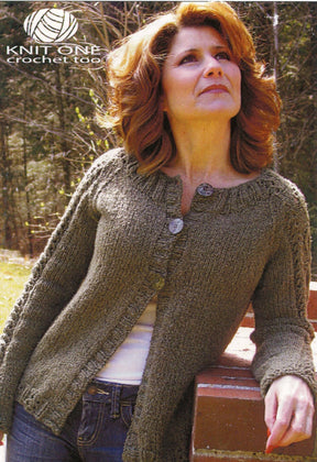 A woman with shoulder-length wavy hair stands outdoors, leaning against a wooden porch rail. She is wearing an Ivy Cardigan by Knit One, Crochet Too in green, featuring three buttons. Underneath, she has on a white top made from worsted weight yarn. In the top left corner of the image are the text "Knit One, Crochet Too" and a yarn logo.