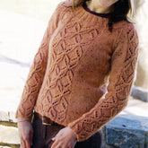 A person wearing the "Winter Butterflies" sweater by Knit One, Crochet Too stands outdoors. The peach-colored sweater showcases intricate leaf and diamond patterns, raglan shaping, a wide ribbed neckline, and long sleeves featuring the same elegant design. The person holds their hands in front of their body.