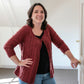 A woman with shoulder-length dark hair, wearing the Vine & Arrows Cardi by Knit One, Crochet Too over a black scoop neck top and blue jeans, stands indoors smiling. Her left hand is on her hip, and her right hand is resting on a doorknob. The background is a brightly lit room with a slanted ceiling.