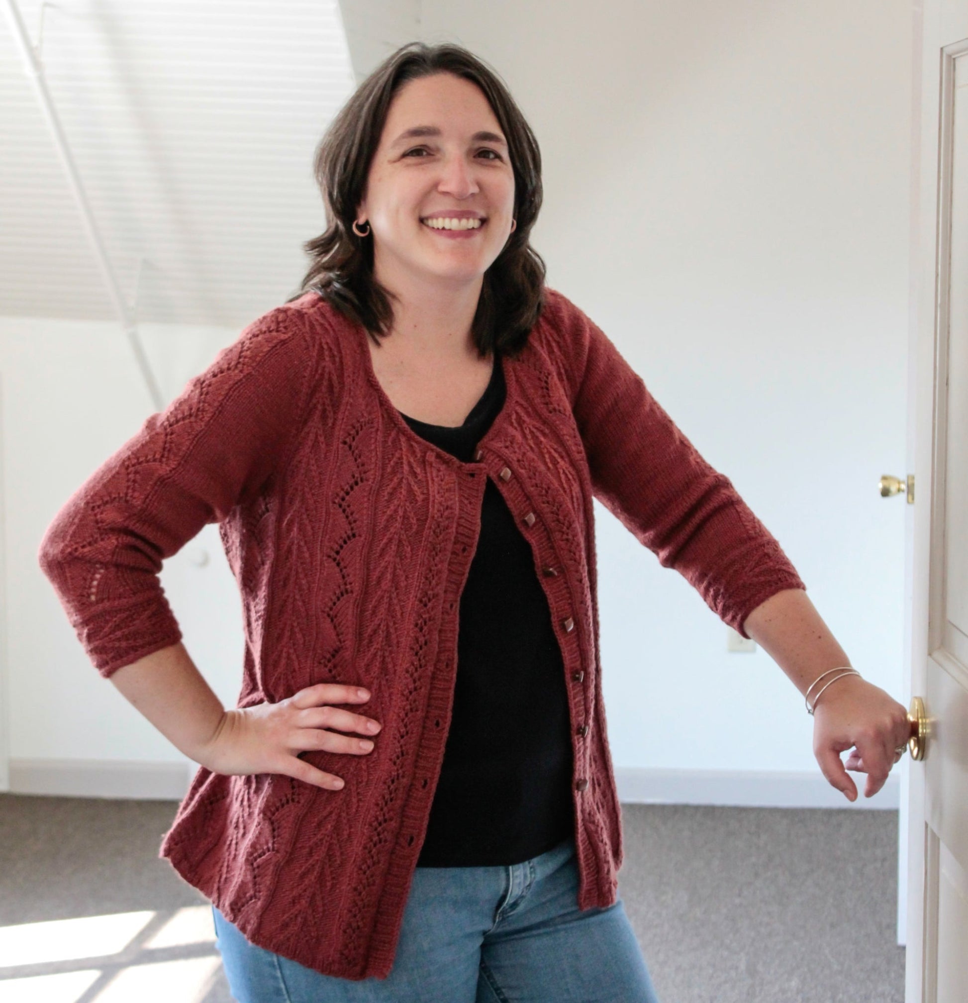 A woman with shoulder-length dark hair, wearing the Vine & Arrows Cardi by Knit One, Crochet Too over a black scoop neck top and blue jeans, stands indoors smiling. Her left hand is on her hip, and her right hand is resting on a doorknob. The background is a brightly lit room with a slanted ceiling.