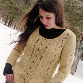 A woman with long brown hair is standing in a snowy outdoor setting, wearing the yellow Vine & Arrows Cardi from Knit One, Crochet Too, over a black scoop neck top. Snow-covered trees appear in the background.