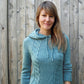 A woman with long, light brown hair stands in front of a wooden fence. She is wearing the Faux Cable Hoodie from Knit One, Crochet Too, adding to its feminine charm. She is smiling gently at the camera.