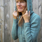 A woman with long hair stands in front of a wooden fence, wearing the Faux Cable Hoodie by Knit One, Crochet Too. The light blue knitted sweater features faux cables and a hood, which she is holding with both hands while smiling slightly and looking up to the side.