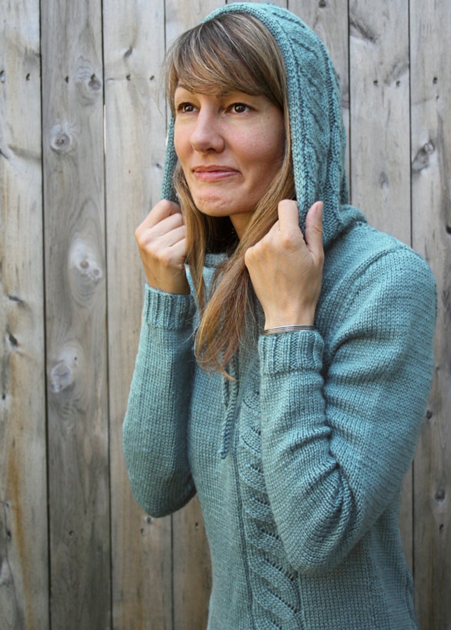 A woman with long hair stands in front of a wooden fence, wearing the Faux Cable Hoodie by Knit One, Crochet Too. The light blue knitted sweater features faux cables and a hood, which she is holding with both hands while smiling slightly and looking up to the side.