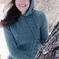 A woman stands outdoors in a snowy forest, smiling and wearing the Knit One, Crochet Too Faux Cable Hoodie in teal. She is leaning against a tree trunk. The text "Knit One, Crochet Too" is displayed in the top right corner.