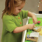 A young child, dressed in a Children's Henley Top Down Sweater from Bryson Distributing, Inc., knit from cozy worsted weight yarn, is playing with a small dollhouse. She arranges miniature beds and dolls, deeply focused on the activity. The dollhouse is white with a green interior and contains tiny furniture and decorations.