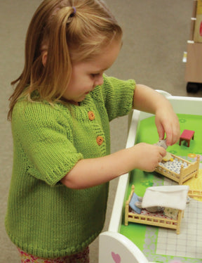 A young child, dressed in a Children's Henley Top Down Sweater from Bryson Distributing, Inc., knit from cozy worsted weight yarn, is playing with a small dollhouse. She arranges miniature beds and dolls, deeply focused on the activity. The dollhouse is white with a green interior and contains tiny furniture and decorations.