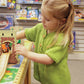 A young girl with blond hair, dressed in a green Children's Henley Top Down Sweater from Bryson Distributing, Inc., plays with a toy train on a wooden track set up on a table. She stands in a store aisle lined with shelves filled with various colorful boxes and toys in the background.