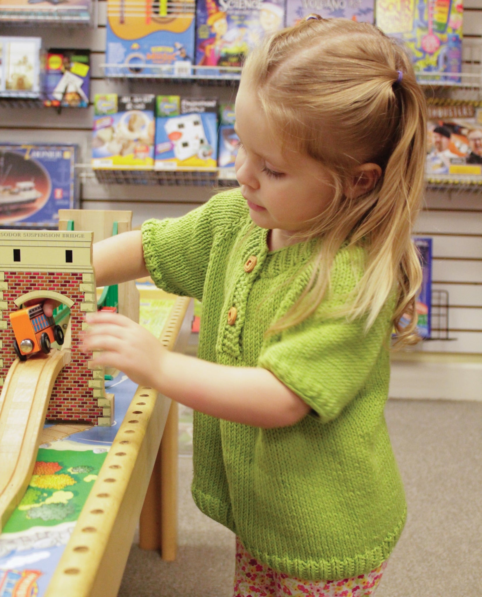 A young girl with blond hair, dressed in a green Children's Henley Top Down Sweater from Bryson Distributing, Inc., plays with a toy train on a wooden track set up on a table. She stands in a store aisle lined with shelves filled with various colorful boxes and toys in the background.