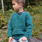 A young boy with short hair wearing a teal Children's Henley Top Down Sweater from Bryson Distributing, Inc., made of worsted weight yarn and light blue jeans sits on a rock in a forested area. He looks to his left, surrounded by greenery including trees and bushes. The setting appears calm and natural.