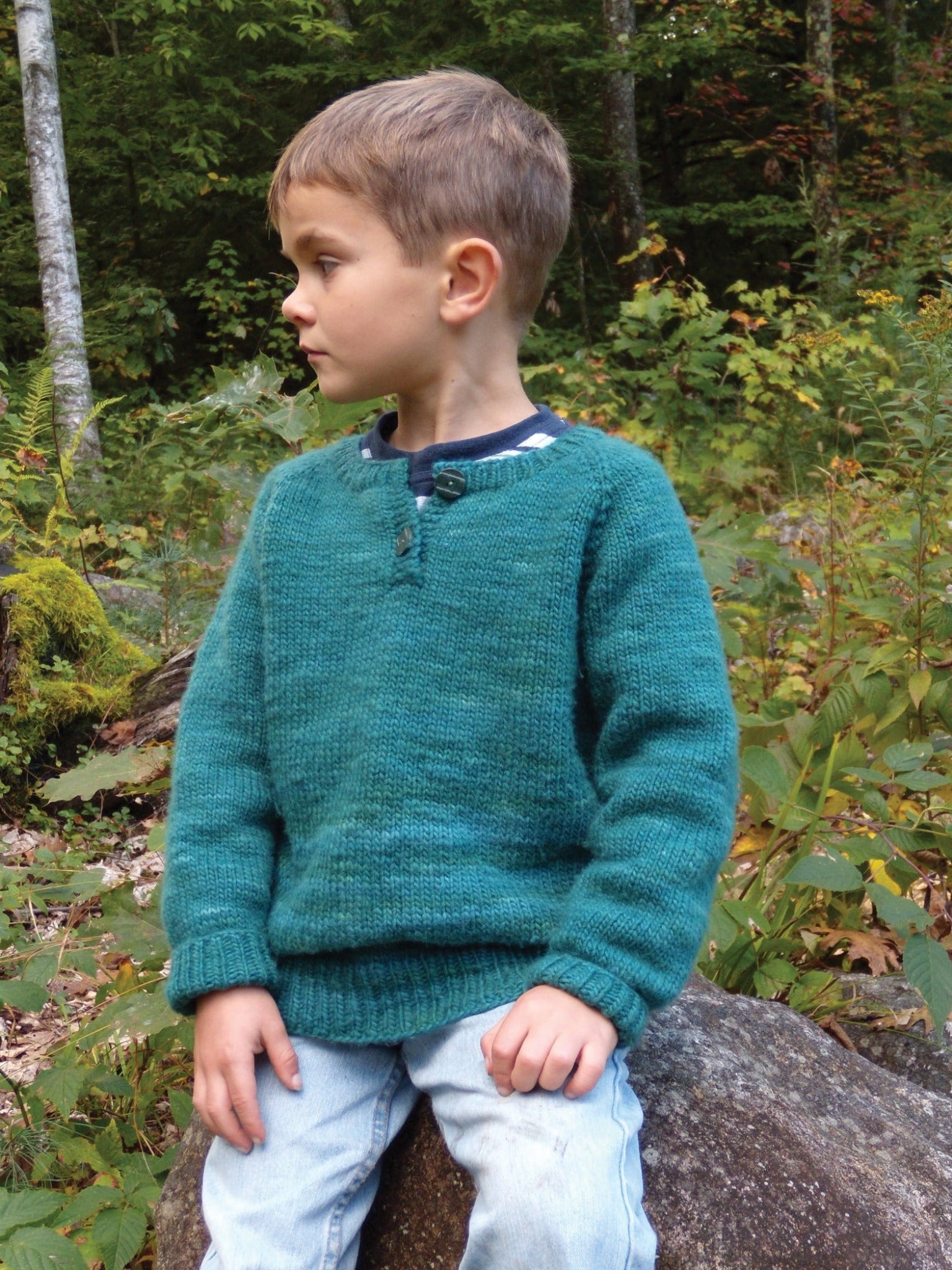 A young boy with short hair wearing a teal Children's Henley Top Down Sweater from Bryson Distributing, Inc., made of worsted weight yarn and light blue jeans sits on a rock in a forested area. He looks to his left, surrounded by greenery including trees and bushes. The setting appears calm and natural.