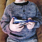 A child sits on a couch, wearing a blue Children's Henley Top Down Sweater by Bryson Distributing, Inc., made of worsted weight yarn. Only the child's torso and hands are visible as they hold a toy model of a predominantly blue U.S. Air Force jet with "U.S. Air Force" written on the side.