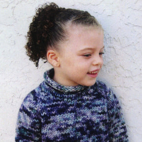 A young child with curly hair tied back into a ponytail, wearing the Children's Bulky Top Down Pullover by Bryson Distributing, Inc., knitted with blue, purple, and white patterns using bulky weight yarn, smiles while looking to the side. The background is a plain, textured white wall.