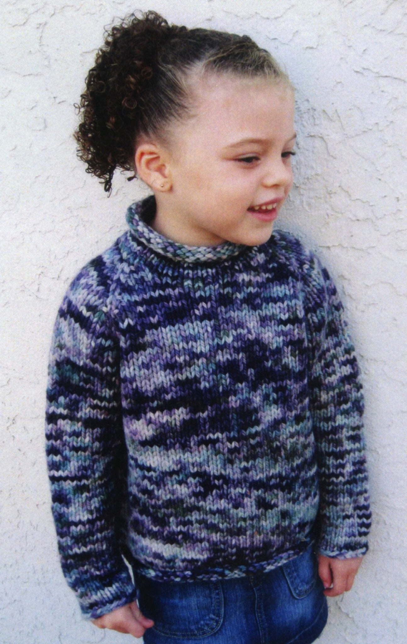 A young girl stands against a textured white wall, smiling and looking to her left. She has curly hair tied up in a ponytail and is wearing Bryson Distributing, Inc.'s Children's Bulky Top Down Pullover that showcases shades of blue, purple, and gray, paired with denim jeans.