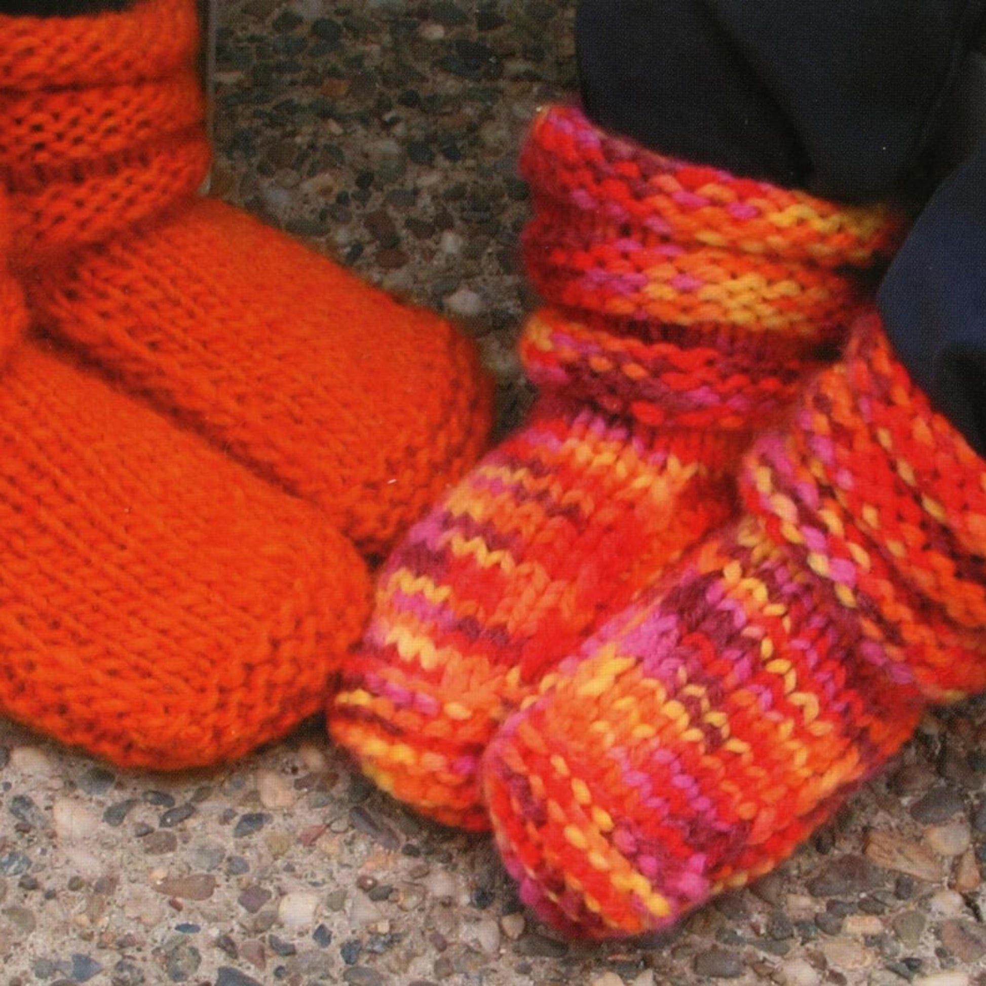 Two pairs of feet adorned with colorful, warm knit Children's Mukluk Slippers from Bryson Distributing, Inc. One pair wears bright orange slippers, while the other sports multicolored slippers featuring shades of red, yellow, purple, and orange. These cozy children's slippers are standing on a pebble-textured surface.