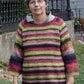 A person with short gray hair and glasses stands outdoors, wearing the colorful, striped Top Down Swing Pullover from Bryson Distributing, Inc. They pose with their hands by their sides. A wrought iron fence and greenery are visible in the background.