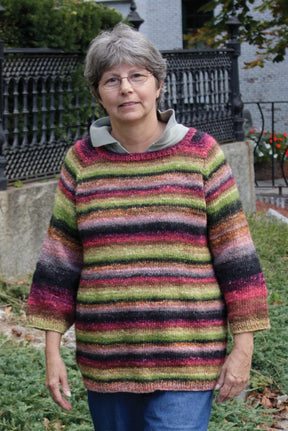 A person with short gray hair and glasses stands outdoors, wearing the colorful, striped Top Down Swing Pullover from Bryson Distributing, Inc. They pose with their hands by their sides. A wrought iron fence and greenery are visible in the background.