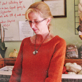 A woman with blonde hair tied back, wearing glasses and an orange sweater, stands in a yarn store. Behind her are wool products on shelves and a handwritten sign on a whiteboard. She appears to be focused on a meticulously laid-out pattern for the Top Down Swing Pullover by Bryson Distributing, Inc.