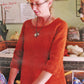 A woman with glasses and blonde hair tied back is wearing the "Top Down Swing Pullover" by Bryson Distributing, Inc., showcasing a comfortable pattern. She is in a room with shelves filled with various yarns and knitting supplies. There is a whiteboard with writing and a certificate on the wall behind her.