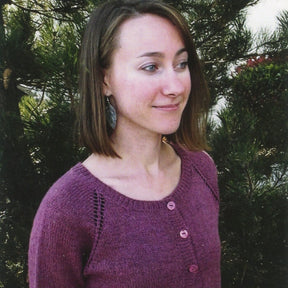 A person with shoulder-length hair stands in front of a tree, wearing Bryson Distributing, Inc.'s Top Down Lightweight Cardigan in purple and an earring shaped like a leaf. The cool evening accentuates the lush green foliage in the background.