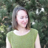 A woman with straight shoulder-length hair and wearing the sleeveless green Side to Side Pullover by Bryson Distributing, Inc., knitted in a garter stitch, stands outdoors in front of dense greenery, smiling softly and looking off to the side.