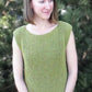 A person with shoulder-length hair stands in front of a tree, wearing the sleeveless Side to Side Pullover by Bryson Distributing, Inc., beautifully showcasing the intricate garter stitch. They are looking slightly to the side, with hands clasped in front. The background is filled with lush green foliage.