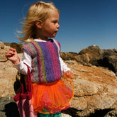 A young child with blonde hair stands on rocky terrain under a clear blue sky. She wears the Side to Side Pullover for Girls by Bryson Distributing, Inc., a colorful, striped knit sleeveless top over her white long-sleeve shirt and an orange tulle skirt. Holding a pink knitted bag, she gazes into the distance, her sweater easy to stretch for playful adventures.